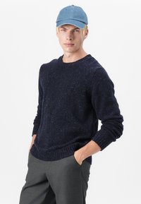 Granatowy sweter dzianinowy o strukturalnym splocie i prążkowanym dekolcie, zestawiony z szarymi, dopasowanymi spodniami i jasnoniebieską czapką z dżinsu.