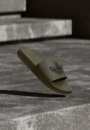 Olivengrøn Adidas slidssandal med sort trekløverlogo, der svæver over teksturerede betontrin under blødt lys.