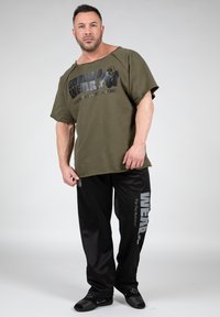 Groene katoenen T-shirt met een grote zwarte graphic, gecombineerd met zwarte joggingsbroek met een zilveren logo-opdruk langs de zijkant. Zwart schoeisel.