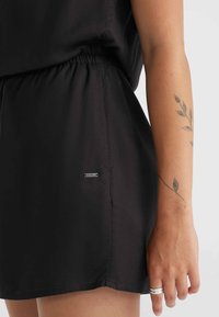 Čierne šortky vyrobené z hladkej tkaniny s elastickým pásom; obsahujú malú kovovú nášivku s logom a jemný lesk.