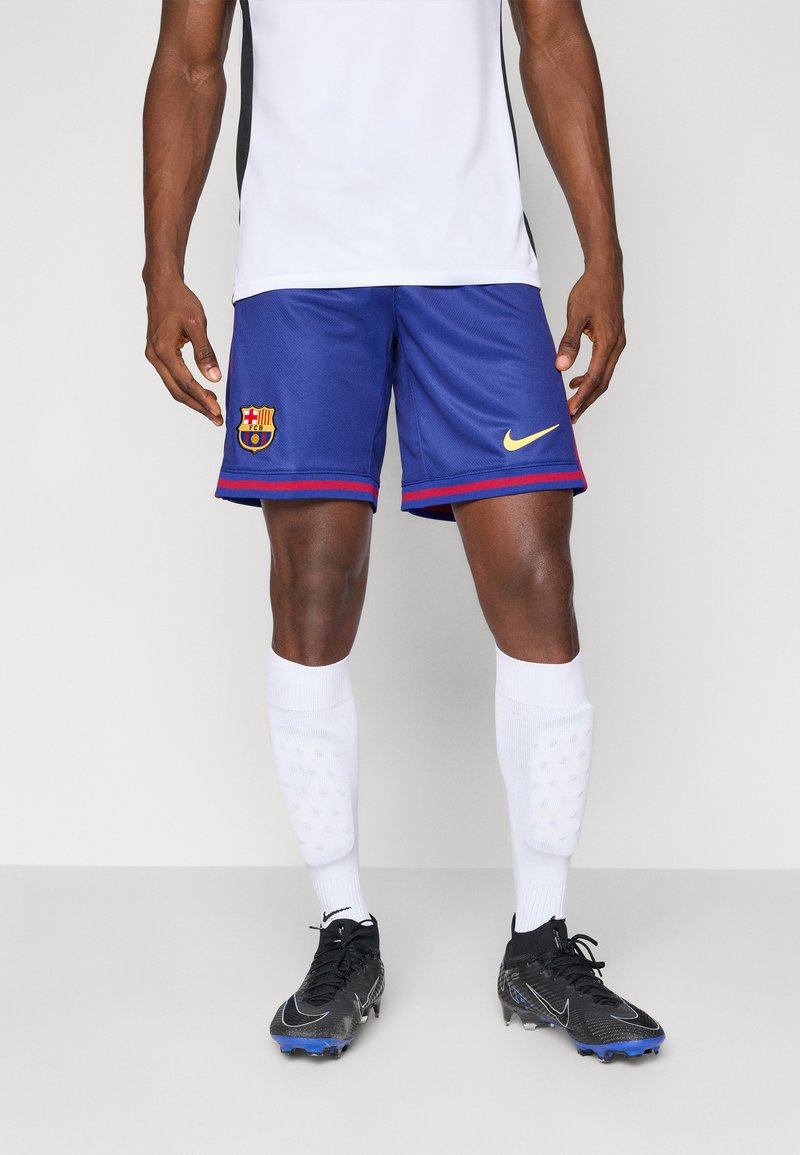 Blue athletic shorts with red accents, Nike logo, and FC Barcelona crest. Worn with white padded socks and black soccer cleats.