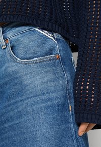 Jeans de mezclilla azul con un ajuste ceñido, que presentan un ligero desgaste, detalles de hardware en color cobre y una etiqueta blanca en el bolsillo. Suéter de punto en color azul marino con textura.