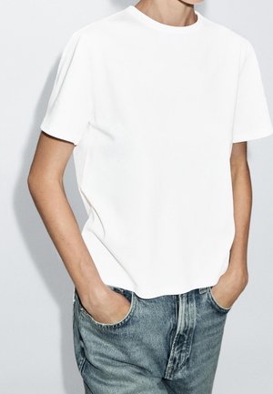 Persoon draagt een effen wit T-shirt met korte mouwen en een blauwe spijkerbroek met de handen in de zakken tegen een effen lichte achtergrond.