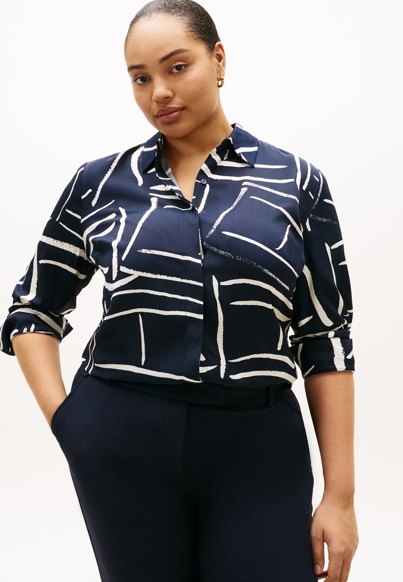 Μπλούζα σε ναυτικό μπλε χρώμα από λείο ύφασμα με άσπρα αφηρημένα γραμμικά σχέδια, διαθέτει γιακά και μανίκια γυρισμένα προς τα πάνω. Συνδυάζεται με παντελόνι σε ναυτικό μπλε χρώμα.