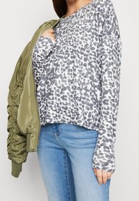 Harmaa ja valkoinen leopardi-printti pitkähihaisessa paidassa, yhdistettynä oliivinvihreään bomber-takkiin, kerroksittain vaaleansinisten farkkujen päällä. Materiaalin tekstuurivaihtelut näkyvissä.