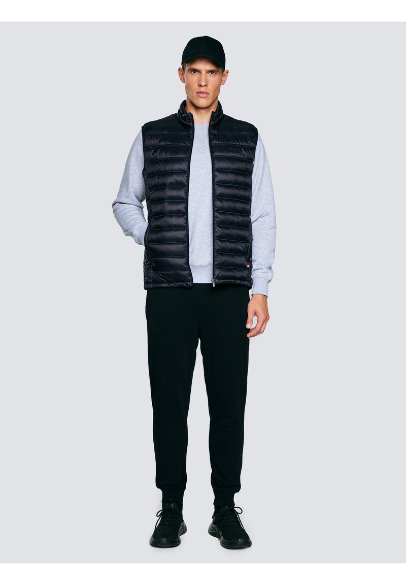 Fiatal férfi fekete sapkában, fekete steppelt mellényben, szürke pulóverben, fekete melegítőnadrágban és fekete sportcipőben, egy kézzel zsebre téve áll.