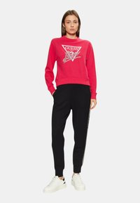 Sudadera roja con el logo "GUESS" en plateado, combinada con pantalones de chándal negros que tienen una franja lateral y zapatillas blancas. Ropa atlética casual.