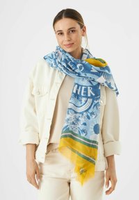 Étole florale bleue et jaune avec une texture légère, arborant des motifs imprimés audacieux et des bords frangés. Portée de manière décontractée autour du cou.