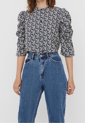 Femme portant un chemisier à manches ballons fleuri bleu marine, rentré dans un jean en denim bleu taille haute, debout devant un fond uni.