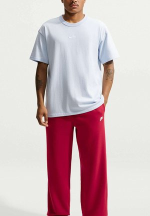 Hombre con camiseta Nike azul claro y pantalones deportivos Nike rojos, de pie con una mano en el bolsillo frente a un fondo liso.