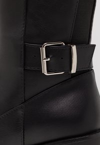 Zbliżenie czarnego skórzanego buta, pokazujące srebrny metalowy klamrę i detale paska z teksturowanym przeszyciem oraz elastycznym panelem.