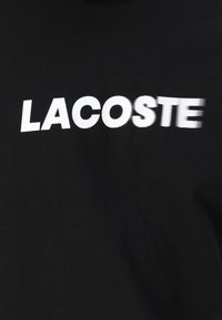 Juodas medvilnės marškinėliai su ryškiu baltu "LACOSTE" logotipu. Dizainas turi glotnią tekstūrą su šiek tiek nusidėvėjusia šrifto išvaizda.