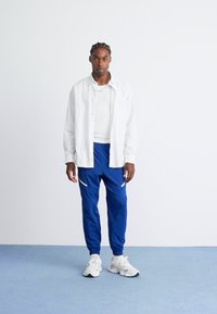 Mand med fletning iført en hvidstribet skjorte over en hvid t-shirt, blå joggers og hvide sneakers, stående på et blåt gulv mod en hvid væg.