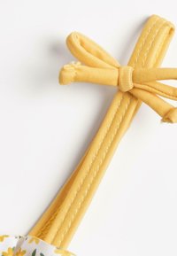 Sangle élastique jaune avec une surface texturée et un accent en forme de nœud. La sangle présente des détails cousus et un motif floral au point de fixation.