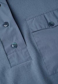 Dettaglio ravvicinato di un tessuto a maglia blu con bottoni blu attaccati e dettagli di tasche a ribalta su un indumento.