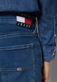 Farkut sinisellä pesulla, joissa on tuettu takataskussa "TOMMY JEANS" -teksti ja punavalko-sinisiä värikorostuksia.