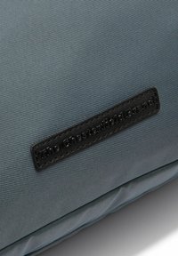 Grauer Stoffkissen mit gerippter Textur, versehen mit einem schwarzen Lederetikett, das mit "The Chesterfield Brand" geprägt ist.