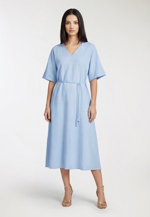 Donna con capelli lisci e scuri che indossa un vestito midi azzurro chiaro con scollo a V, maniche corte e cintura annodata, in piedi davanti a uno sfondo bianco semplice.