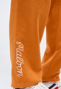 Close-up of orange sweatpants with white cursive "tealer" text embroidered near the ankle, paired with white sneakers with navy and red stripes.