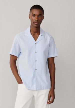 Homme portant une chemise à manches courtes bleu clair à boutons marron et un pantalon blanc, debout avec une main dans la poche.