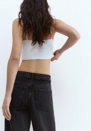 Frau mit langen dunklen Haaren trägt ein hellgraues, bauchfreies Tanktop und hoch taillierte schwarze Jeans und steht vor einem schlichten weißen Hintergrund.