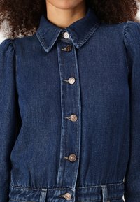 Mørkeblå denimjakke med krage, puffede lange ærmer og brune knapdetaljer langs fronten og taljen.