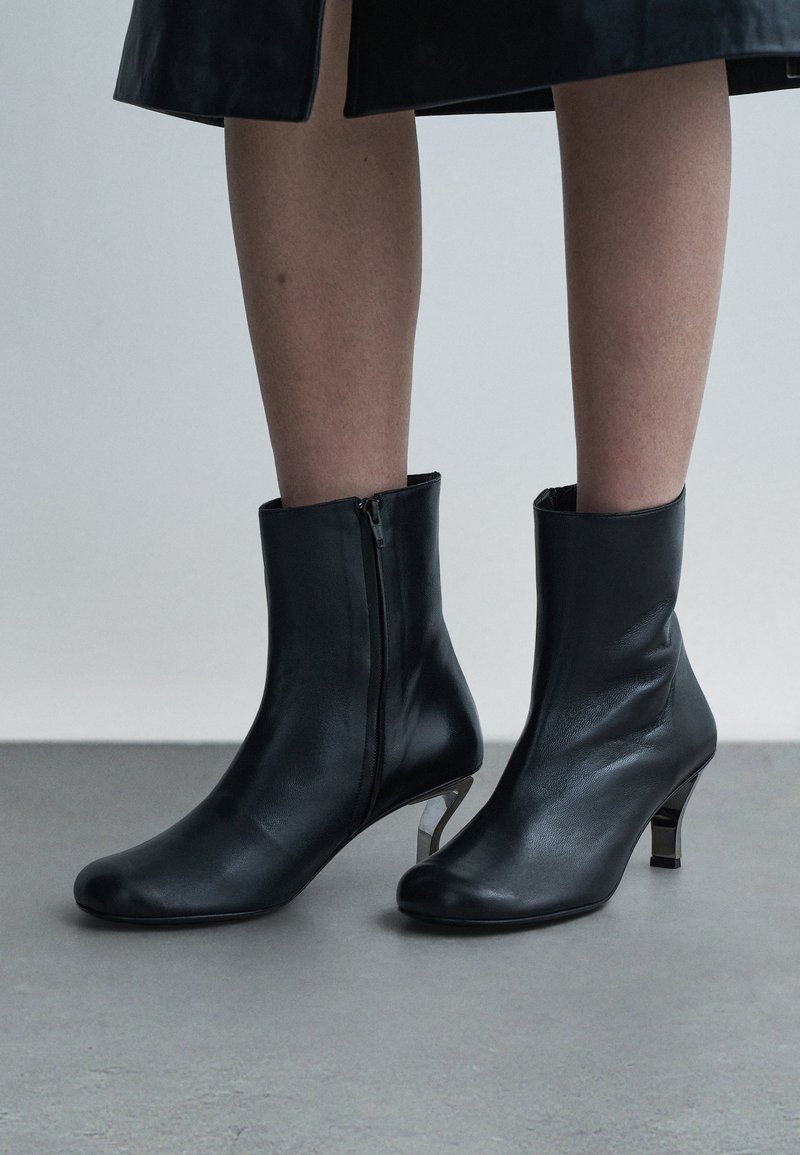 Black ankle boots made of smooth leather with a side zipper, featuring a short geometric heel and a sleek, minimalist design.