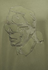 Αφηρημένη κεντημένη περίγραμμα άνδρα που φορά γυαλιά ηλίου και μπουφάν με ψηλό γιακά σε ελιά πράσινο ύφασμα.