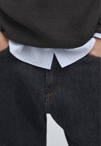 Ciemnoszary sweter z dzianiny nałożony na jasnoniebieską koszulę w paski z kołnierzykiem, schowaną w ciemne dżinsy z pomarańczowym przeszyciem.