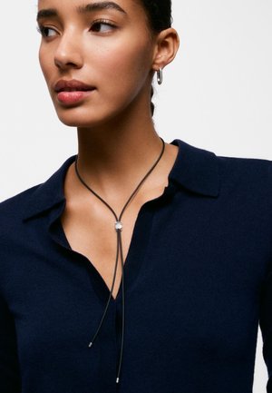 Kobieta z zaplecionymi włosami nosi małe srebrne kolczyki-koła i granatową bluzkę z kołnierzykiem, która ma czarny skórzany naszyjnik bolo z srebrną klamrą.