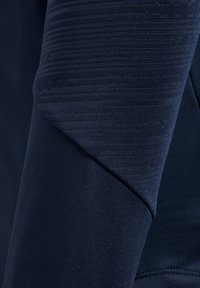 Gros plan d'une manche en tissu bleu foncé avec des sections tricotées lisses et texturées formant un motif géométrique.