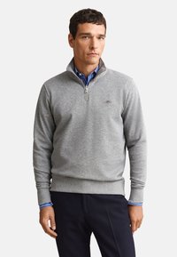 Homme portant un pull gris à fermeture éclair quart de zip sur une chemise bleue rayée et un pantalon sombre, debout avec une expression neutre sur un fond uni.