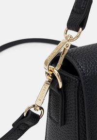 Närbild av strapped läderväska i svart med textur, fäst med gyllene metallklämmor graverade med "VALENTINO"-logotypen.