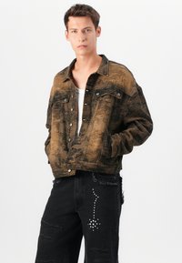 Chaqueta de mezclilla desgastada en marrón y negro con cierre de botones, bolsillos en el pecho y corte relajado. Combinada con jeans negros rasgados que presentan tachuelas.