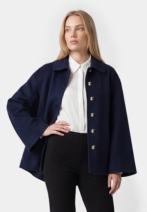 Kvinde med glat blondt hår har en mørkeblå jakke med knapper over en hvid skjorte med krave og sorte bukser på, poserer mod en ensfarvet baggrund.