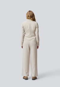 Ljust beige kostym med en skräddarsydd kavaj och vida byxor. Tyget verkar ha en texturerad yta med knappar längs ärmarna och en figursydd midja.