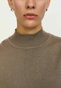 Jednostavna smeđa visok vratna džemper s rebrastom teksturom. Blizak prikaz naglašava dizajn vrata i glatku kvalitetu tkanine. Nema vidljivih uzoraka ili ukrasa.
