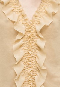 Ύφασμα σε κρεμ απόχρωση με συσσωρευμένες και στρωματοποιημένες βολάν που δημιουργούν ένα κατακόρυφο διακοσμητικό στοιχείο σε ρούχο.