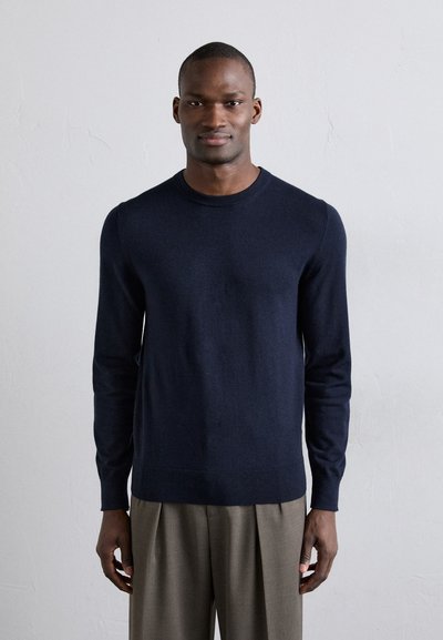 Maglione a maglia blu navy con collo rotondo, maniche lunghe, trama fine e polsini a coste. Abbinato a pantaloni grigi chiari plissettati.