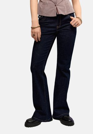 Osoba nosiaca tmavomodré bootcut rifle, čierne robustné topánky, kockovanú košeľu a zlaté náramky a prstene na oboch rukách.