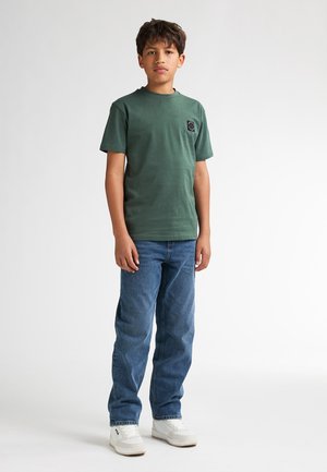 Groen katoenen T-shirt met een ronde hals en een klein zwart logopatch, gecombineerd met blauwe spijkerbroek en witte sneakers.