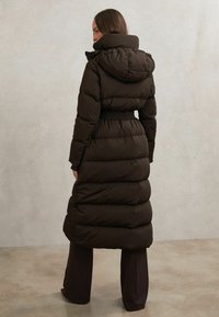 Piumino lungo marrone scuro con vita cinturata, collo alto e cappuccio. Realizzato in tessuto trapuntato con una texture liscia e imbottita.