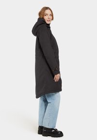 Schwarzer langer Mantel mit Kapuze, aus glattem Material. Mit seitlichen Taschen und geknöpften Bündchen. Getragen über hellblauen Jeans und schwarzen Stiefeln.