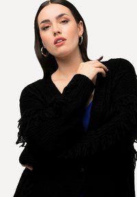 Cardigan nero lavorato a maglia con dettagli a frangia sulle maniche, caratterizzato da una scollatura a V profonda e una sottile texture a coste. Indossato sopra una maglietta blu.