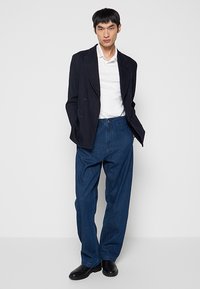 Granatowa marynarka dwurzędowa na białej koszuli, połączona z luźnymi, ciemnoniebieskimi dżinsami i czarnymi butami. Prosty i uporządkowany design.