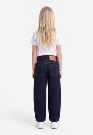 Kind met lang blond haar staat met de rug naar de camera, draagt een wit t-shirt, donkere wijde jeans en witte sneakers tegen een witte achtergrond.