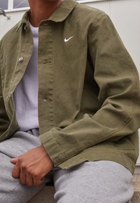 Vihreä kevytrakenteinen takki, jossa on napitettava etuosa ja valkoinen Nike-logo. Päälle puetun harmaan t-paidan ja harmaiden hupparihousujen kanssa.