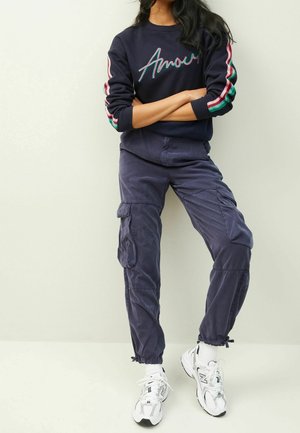 Osoba stojí se založenýma rukama, oblečená v tmavě modré mikině s nápisem "Amour", tmavých cargo kalhotách a bílých teniskách na jednobarevném pozadí.