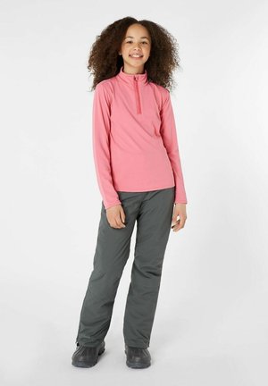 Langemouwen, roze top met ritssluiting, gecombineerd met donkergrijze broek. De outfit is ontworpen voor comfort en buitenactiviteiten. Zwarte laarzen maken de look compleet.