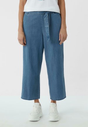 Kvinde iført blå vidde jeansbukser med bindebånd foran og hvide plateau-sneakers, stående mod en ensfarvet baggrund.